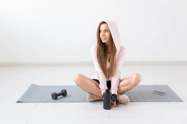 Fitness, deporte, concepto de la gente - mujer joven está sentada en la alfombra después de un ejercicio y mirando hacia los lados, relajándose —  Fotos de Stock