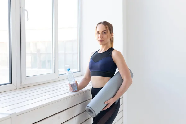 Gente, deporte y fitness concepto - mujer joven con esterilla de yoga y botella de agua de pie cerca de la ventana —  Fotos de Stock