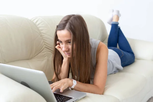 Технологии, фрилансер и концепция людей - красивая молодая женщина лежит на диване с ноутбуком — стоковое фото