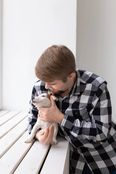 people, pets and animals concept - young man hugging jack russell terrier puppy near window on white background