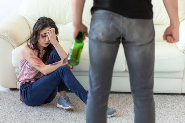 Victim, domestic violence, abuse and alcoholic concept - drunk man with bottle near his wife sitting on the floor — Stock Photo, Image