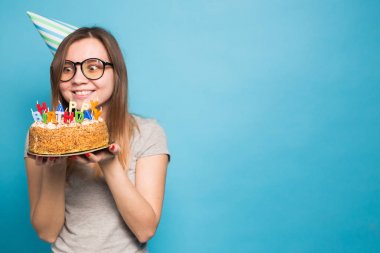 Sevimli neşeli çılgın genç kız öğrenci kutlama kağıt şapka elinde mavi bir arka planda ayakta mutlu bir Doğum günü pastası tutarak. Reklam alanı.