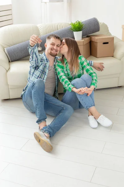 Charming young woman kisses her husband holding in hands the keys to their new apartment while sitting in her new living room