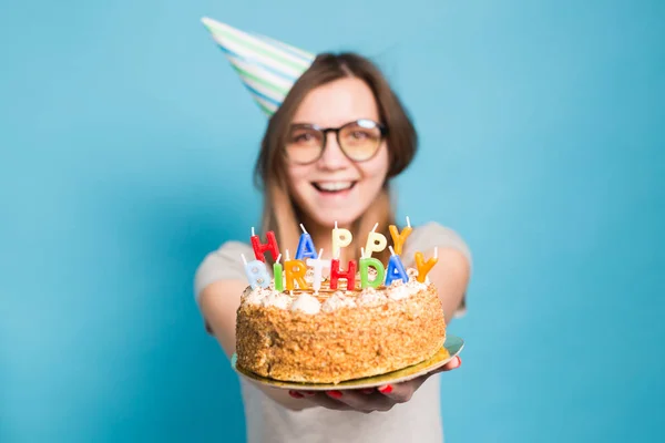 Fechar engraçado desfocado menina positiva em óculos e chapéu de papel saudação segurando um bolo de aniversário feliz em suas mãos em pé sobre um fundo azul . — Fotografia de Stock