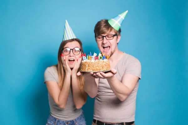 Pareja joven divertida en gorras de papel y con un pastel hacen una cara tonta y desean feliz cumpleaños mientras están de pie sobre un fondo azul. Concepto de felicitaciones y bromas . — Foto de Stock