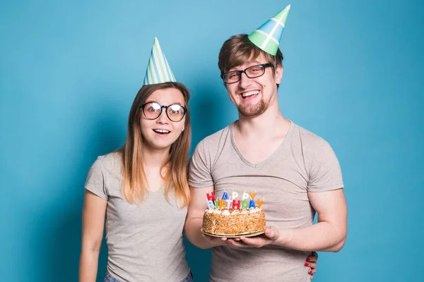 Alegre joven pareja encantador chico y lindo chica en papel sombreros hacer tonto cara y mantener en sus manos un pastel con la inscripción cumpleaños de pie sobre un fondo azul . — Foto de Stock