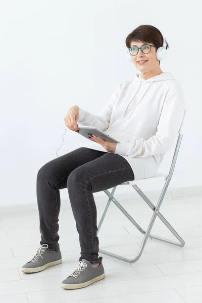 Charming slim middle-aged woman in glasses and a sweater sits on a chair and listens to her favorite online radio with the help of a headset and wireless internet on a white background — Stock Photo, Image