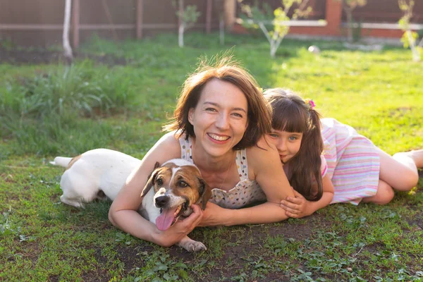Liten flicka i rosa klänning med mor och Jack Russell Terrier hund om på gräs. Titta på kamera och leende — Stockfoto
