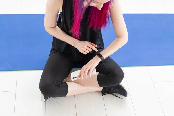 Fiatal vékony azonosítatlan nő, lila hajú keres a pulzus és elégetett kalória segítségével okos óra és egy okostelefon ülve sport szőnyeg egy tornateremben. A sportalkalmazás fogalma. — Stock Fotó