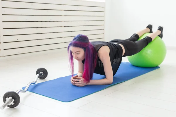 Joven chica asiática con el pelo de color hace un ejercicio de tablón utilizando fitball en una alfombra deportiva azul junto a una barra. Concepto de entrenamiento en el gimnasio y buena forma física . — Foto de Stock