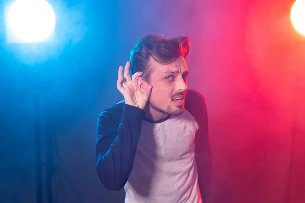 Young embarrassed handsome man hipster with a beard leaned his hand to his ear trying to listen to pose on a red blue background. Concept of importance of opinion. — 스톡 사진