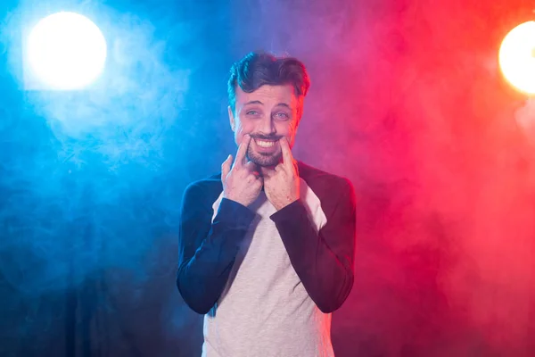 Un joven divertido con barba en ropa casual saca una sonrisa con las manos posando sobre un fondo azul rojo en el estudio. Concepto de ánimo triste e hipocresía . — Foto de Stock