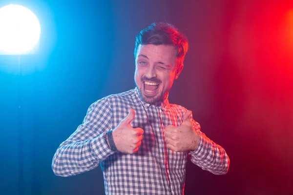 Riendo joven barbudo con una camisa a cuadros que muestra el pulgar hacia arriba posando en el estudio sobre fondo azul-rojo. El concepto de una elección exitosa y el éxito . — Foto de Stock