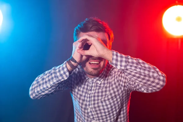 Ο νεαρός άντρας με γένια με τα χέρια του απεικονίζει ένα τηλεσκόπιο που ποζάρει σε κόκκινο-μπλε φόντο. Η έννοια της ανάκτησης πληροφοριών και ο αιτών. — Φωτογραφία Αρχείου
