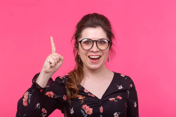 Retrato de una joven y romántica joven estudiante con gafas que muestran su dedo posando sobre un fondo rosa. Espacio para la publicidad. Concepto de nuevas ideas . — Foto de Stock