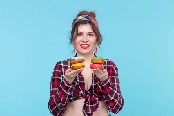 Pozytywny młody pin-up girl model trzyma w rękach cztery wielokolorowe pączki stwarzające na niebieskim tle. Koncepcja gotowania deserów i słodyczy. — Zdjęcie stockowe