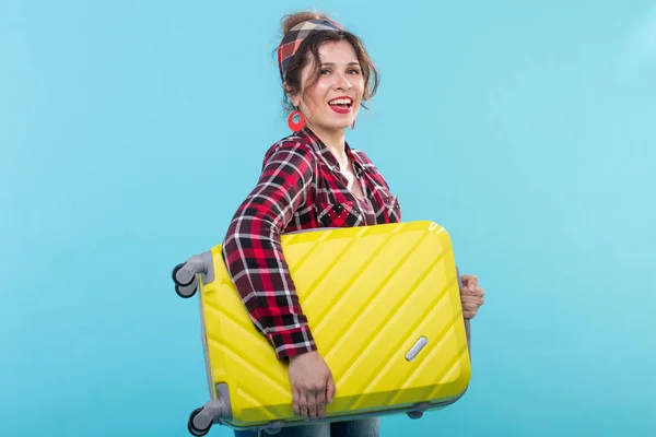 Joven mujer sonriente positiva con una camisa a cuadros que sostiene una maleta amarilla posando sobre un fondo azul. Concepto de turismo y viajes — Foto de Stock