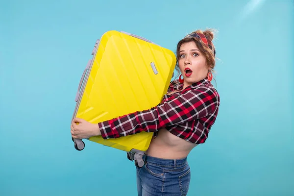 Frightened young beautiful woman in vintage clothes holding a yellow suitcase in her hands posing on a blue background. Concept travel with a heavy load.