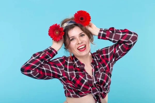 Портрет смішної усміхненої красивої молодої жінки в червоній картатій сорочці, що замінює червоні квіти їй на голову, позуючи на синьому тлі. Концепція святкування та дива . — стокове фото