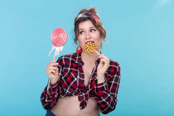 Charming young woman in retro clothes holding colorful lollipops in her hands and licking one posing on blue background. Concept of love for sweets and holidays.