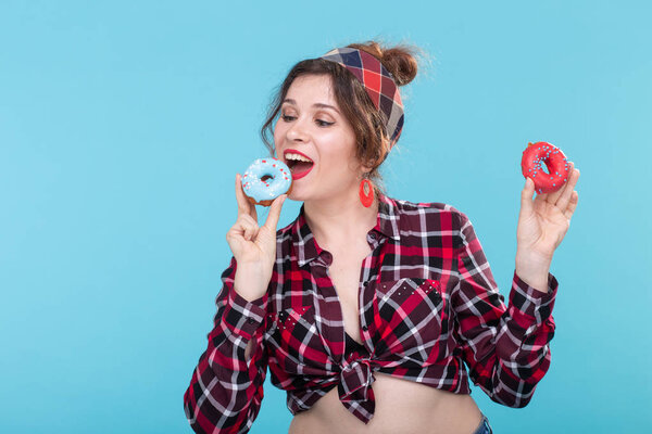 Beautiful young charming woman on vintage clothes wants to bite off blue donut and holds in her hands a red one posing against blue background. Concept of delicious pastries and desserts from cooking.