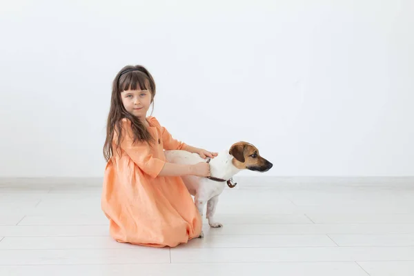 Liten charmig flicka med mörkt hår i en lång klänning sitter bredvid hunden Jack Russell Terrier på golvet i hennes rum. Begreppet barn oskuld och barnomsorg. Copyspace. — Stockfoto
