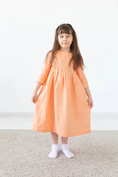 Vacker liten flicka i en persika klänning poserar mot en vit vägg. Begreppet barndesigner kläder. — Stockfoto