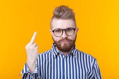 Bıyıklı ve sakallı gözlüklü kızgın, genç ve şık bir adam, sarı bir arka plan üzerinde poz veren orta parmağını gösteriyor. İnkar ve sıkıntı kavramı.