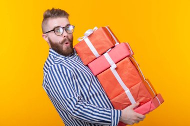 Sarı bir arka planda hediye kutuları büyük bir yığın tutan gözlüklü genç hipster adam şaşırttı. Yeni yıl ve yılbaşı hediyeleri kavramı