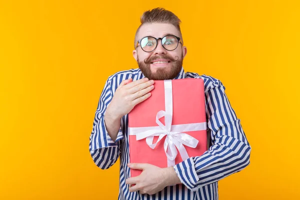 戴着胡子、戴着胡子的快乐年轻时尚男子在黄色背景上拥抱着一个红色的礼品盒。礼物的快乐的概念. — 图库照片