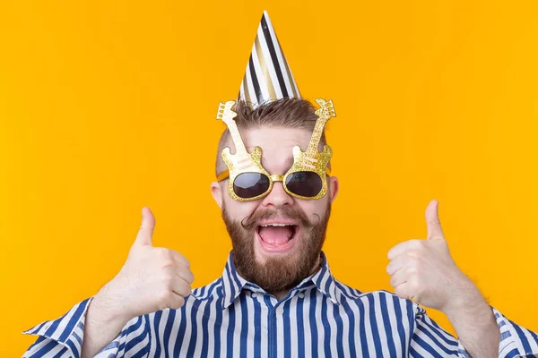 Gek vrolijke vrolijke jonge man in een pet en bril toont duimen omhoog op een gele achtergrond. Het concept van een geslaagde feest en vakantie. — Stockfoto