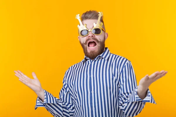 Joven positivo con gafas en forma de guitarras se alegra de haber levantado las manos sobre un fondo amarillo. El concepto de celebración y fiestas . — Foto de Stock