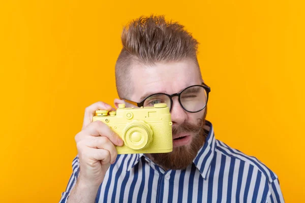 Fotógrafo jovem bonito com bigode e barba está fotografando uma câmera vintage amarela em um fundo amarelo. Conceito de hobby e trabalho profissional . — Fotografia de Stock