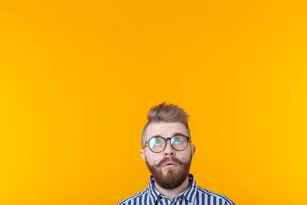 Sorprendido joven hipster hombre con bigote y con gafas posando sobre un fondo amarillo en sorpresa mirando hacia arriba. Concepto de sorpresa y lugar de publicidad y enlaces . — Foto de Stock