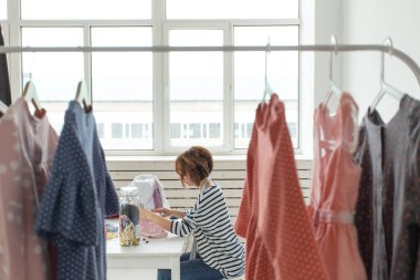 Gözlüklü bir elbise askısı genç terzi kadın görünümü aksesuarları ile masada oturan bir dikiş makinesi üzerinde yeni bir ürün diker. Tasarımcı giysi kavramı.