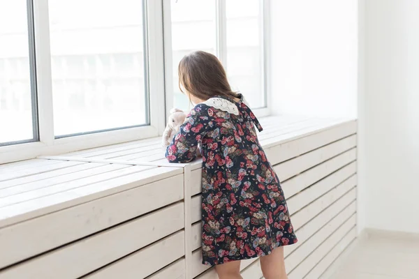 Vue latérale d'une petite fille non identifiée dans une longue robe à fleurs tenant un lapin préféré dans ses mains et regardant par la fenêtre dans sa chambre. Le concept de rêverie enfantine . — Photo