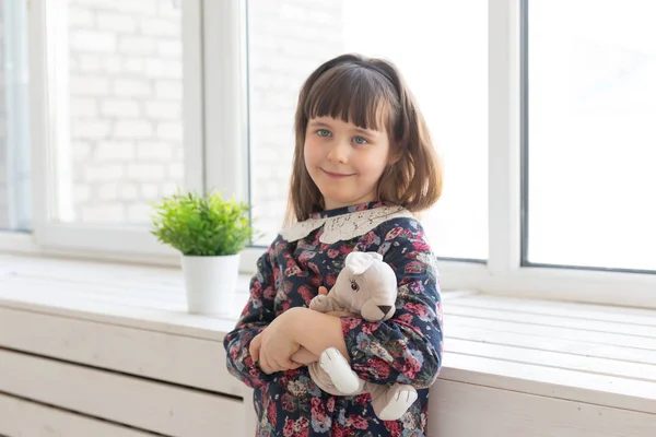 Porträtt av en söt snäll liten flicka i en blomma klänning med en favorit mjuk kanin leker leksak i händerna stående nära fönstret. Konceptet med Childs rum och relaterade produkter. — Stockfoto