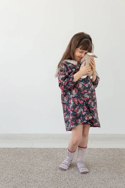 Маленькая очаровательная девочка в цветочном платье нежно обнимает своего любимого игрушечного кролика, стоящего на белом фоне. Концепция детских игрушек и товаров — стоковое фото