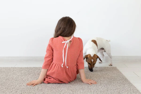 Visão traseira de uma menina em um vestido de bolinhas vermelhas sentado no chão ao lado de seu amado cão Jack Russell Terrier em um fundo branco. O conceito de produtos infantis . — Fotografia de Stock