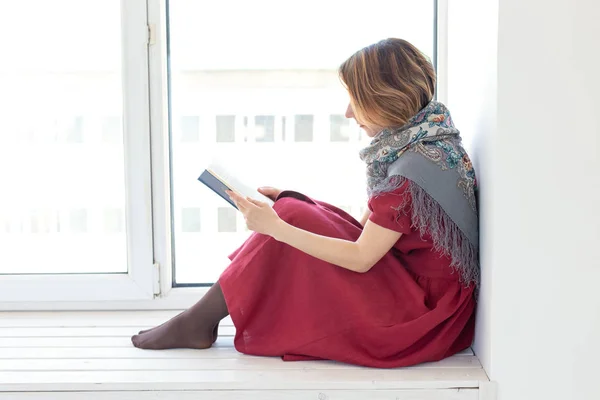 Uzun mütevazı bir elbise ve bir eşarp içinde büyüleyici bir genç kız Yan görünümü pencere üzerinde otururken bir kitap okuyor. Gençlik ve romantizm kavramı. — Stok fotoğraf