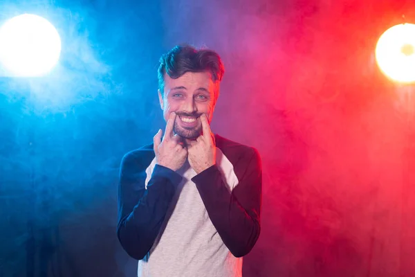 Ein lustiger junger Mann mit Bart in lässiger Kleidung zaubert im Studio ein Lächeln mit seinen Händen, die auf einem rotblauen Hintergrund posieren. Konzept von trauriger Stimmung und Heuchelei. — Stockfoto