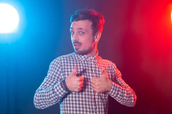 Joven guapo con barba hipster en una camisa a cuadros posando sobre un fondo azul-rojo y mostrando los pulgares hacia arriba. Concepto de éxito y buena elección . — Foto de Stock