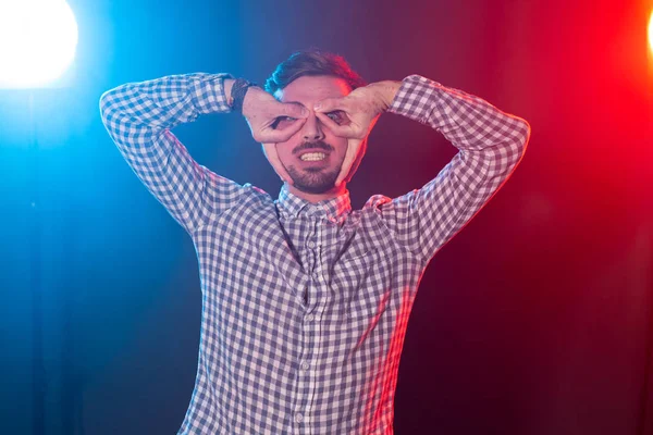 Chico hipster joven positivo divertido en una camisa a cuadros bromas mientras que representa sus gafas sobre un fondo azul-rojo . — Foto de Stock