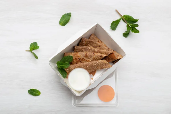 Pfannkuchen mit Sauerrahmsoße in einer weißen Lunchbox liegen auf einem weißen Tisch neben dem Grün. Ernährungskonzept für Snacks. — Stockfoto