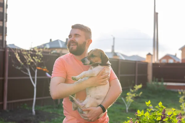 Jovem positivo com uma barba está segurando seu amado cão Jack Russell Terrier e está desfrutando de um descanso conjunto no quintal de sua casa de campo em um dia ensolarado de verão. O conceito de amor para os seus quatro — Fotografia de Stock