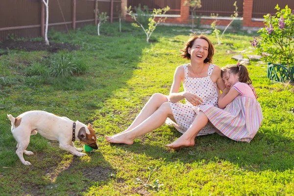 Söt ung mor och dotter sitta på gräset på sitt hus barfota bredvid sin älskade hund på en solig sommardag. Familjesemester koncept. — Stockfoto