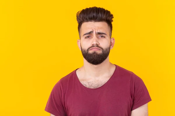 Triste jovem e bonito hipster com uma barba em uma camiseta borgonha posando em um fundo amarelo. Conceito de frustração . — Fotografia de Stock