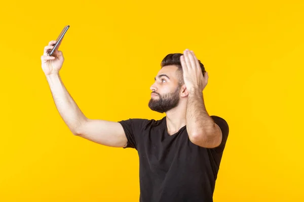 Hipster bonito jovem barbudo tomando uma selfie em seu smartphone sobre fundo amarelo. Conceito de narcisismo nas redes sociais . — Fotografia de Stock