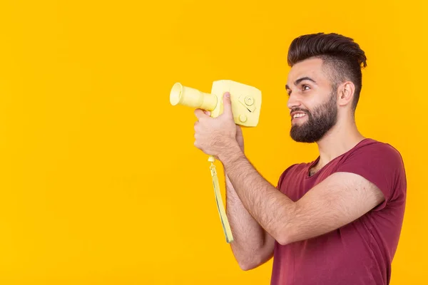 Side view of a handsome young male hipster with a beard shoots objects on a vintage video camera on a yellow background with copy space. Concept of video — Stock Photo, Image