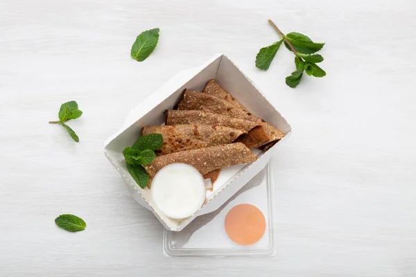 Vue de dessus des crêpes à la crème sure dans une boîte à lunch blanche se trouvent sur une table blanche à côté des verts. Concept de collation diététique . — Photo